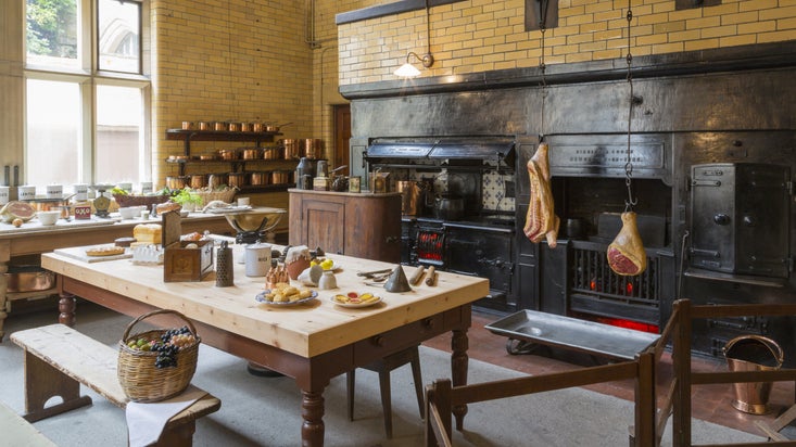 The Kitchen in Cragside House with buttery yellow glazed brick walls, a grand wooden work table, double range and rotating roasting spit.